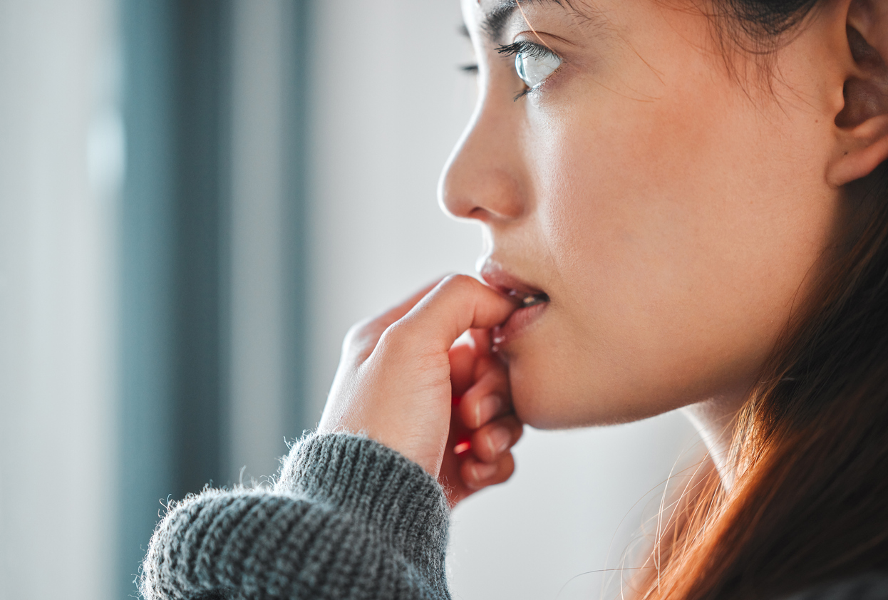 A closeup of a woman with anxiety biting her thumb while looking out a window