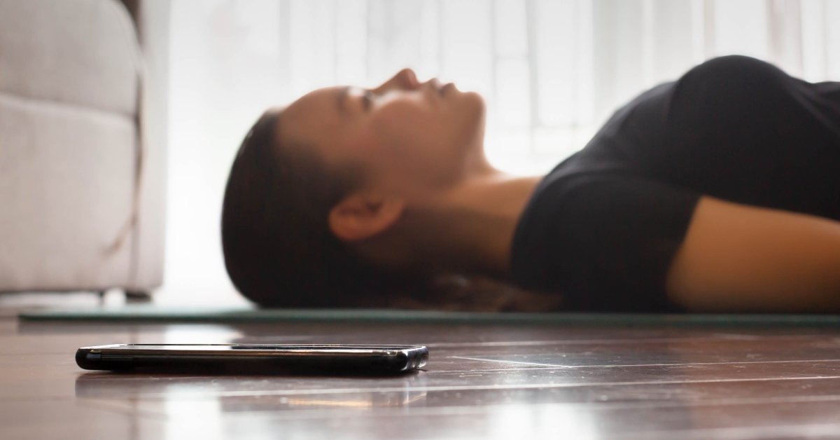 A woman is lying on the floor with her eyes closed. A smart phone lies on the floor in the foreground.