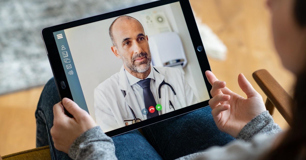 A woman sitting on a couch looking at a tablet. A doctor with a stethoscope is visible on the screen.