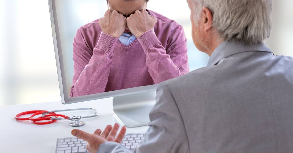 Older doctor sitting at a computer. The young man on the screen seems down and is resting his face in his balled-up fists.