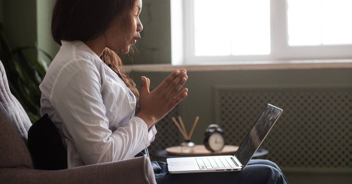 Young professional woman sitting in a chair with her hands clasped together. Sitting on her knees is an open laptop.
