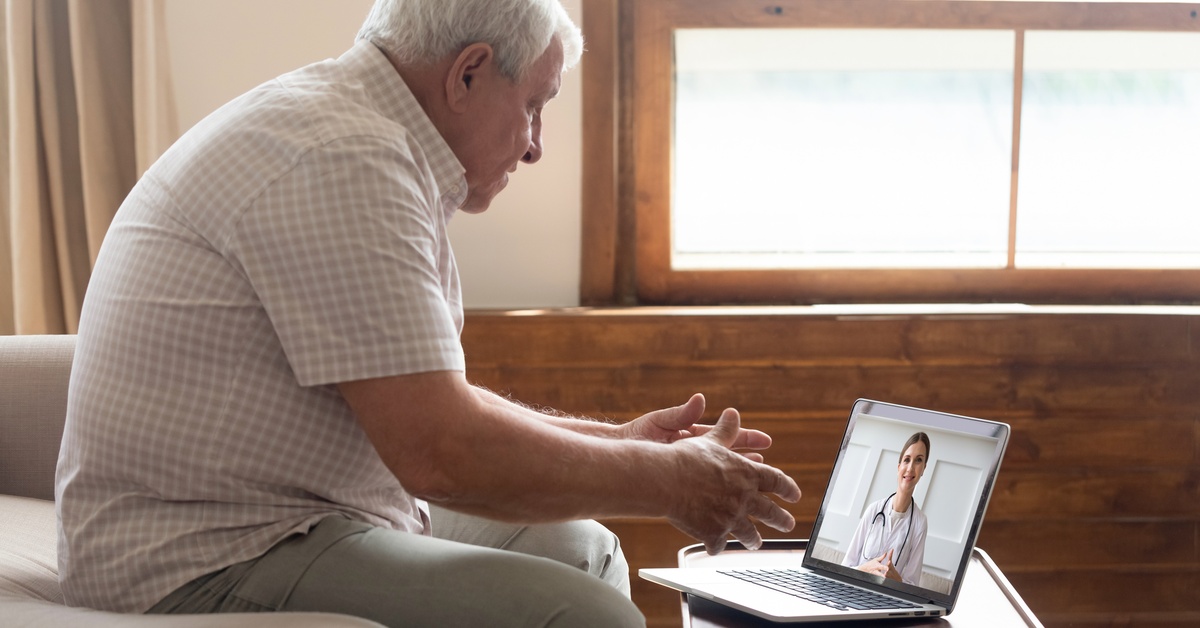 An older man on a sofa is talking to a doctor on a laptop, which is on a coffee table. The doctor on the screen has her hands folded.