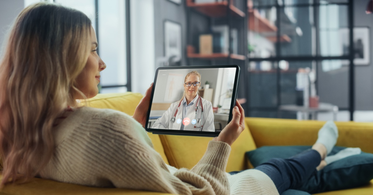 A woman lying on a yellow couch talking to a female doctor on the screen of a tablet she's holding.