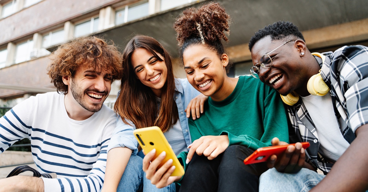 A group of young people sit outside together and laugh while staring at the contents of a smartphone.
