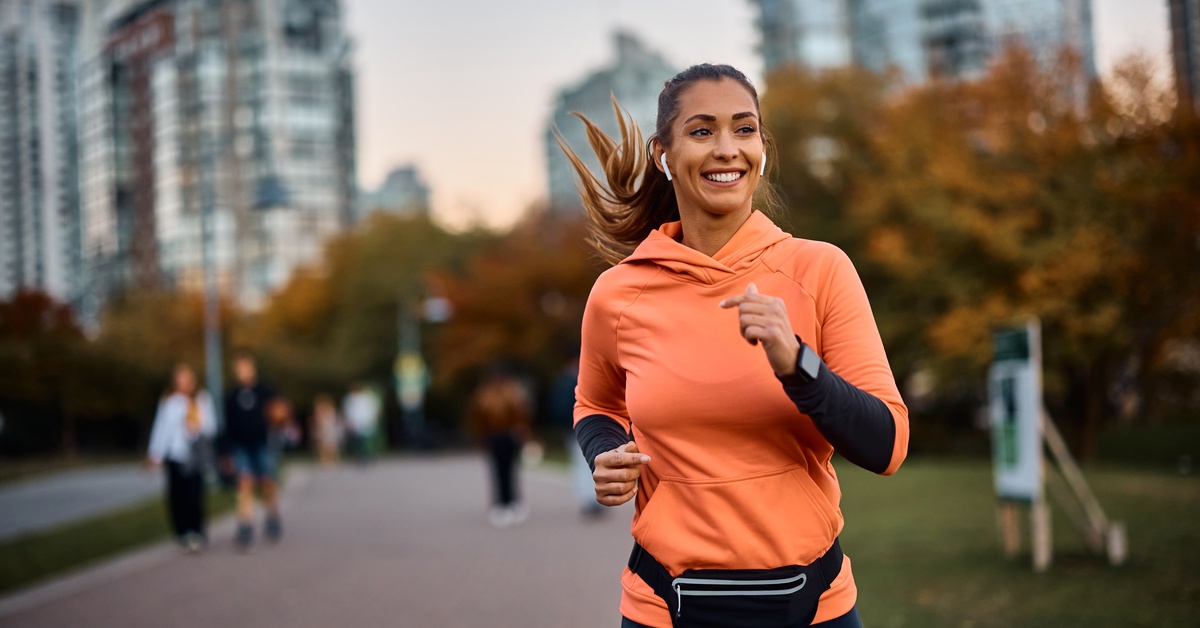 A young woman wearing an orange sweatshirt, black fanny pack, and white earbuds smiles while running in a park.