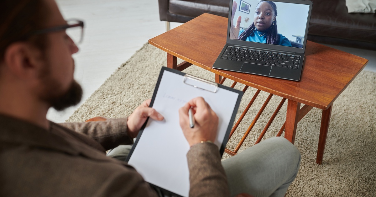 A man with glasses and a clipboard is speaking to a woman on the screen of a laptop on his coffee table.