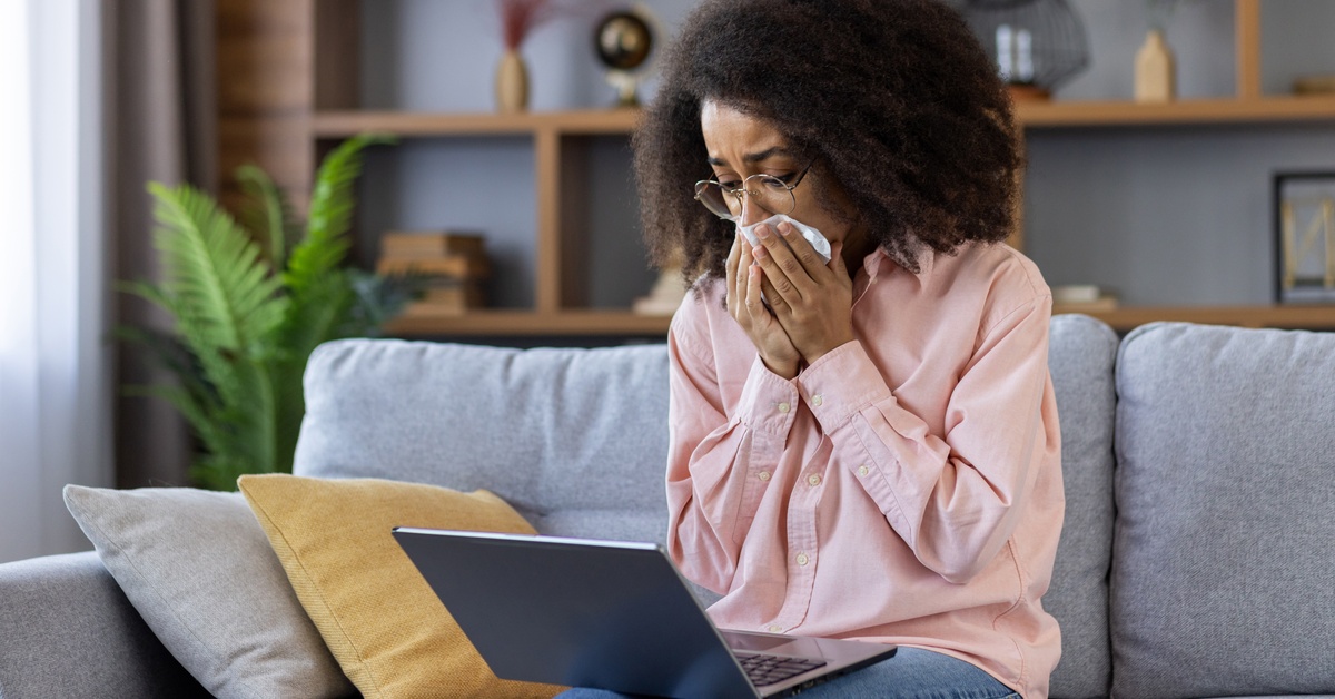 A young woman in a pink sweatshirt is sitting on a couch and holding a tissue up to her mouth. A laptop rests on her knees.