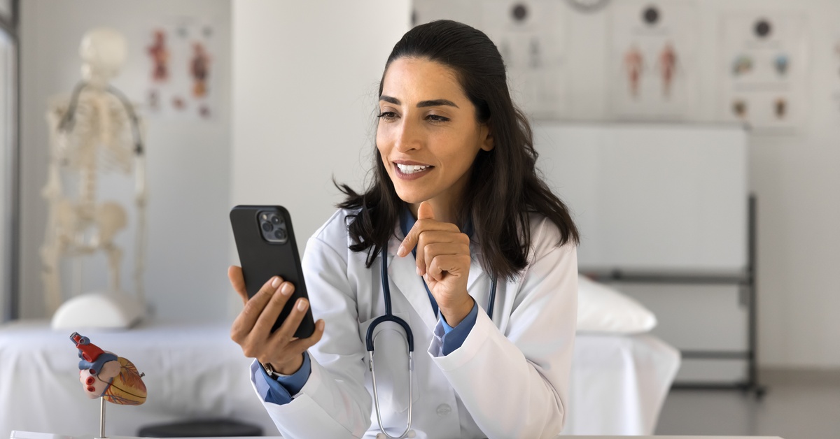 A female doctor wearing a white lab coat and stethoscope uses a smartphone to conduct an appointment in a medical office.