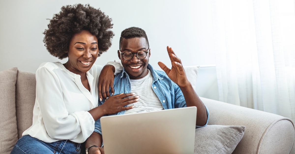 A man and a woman sit close together on a couch, looking at a laptop screen. They each have a smile on their face.