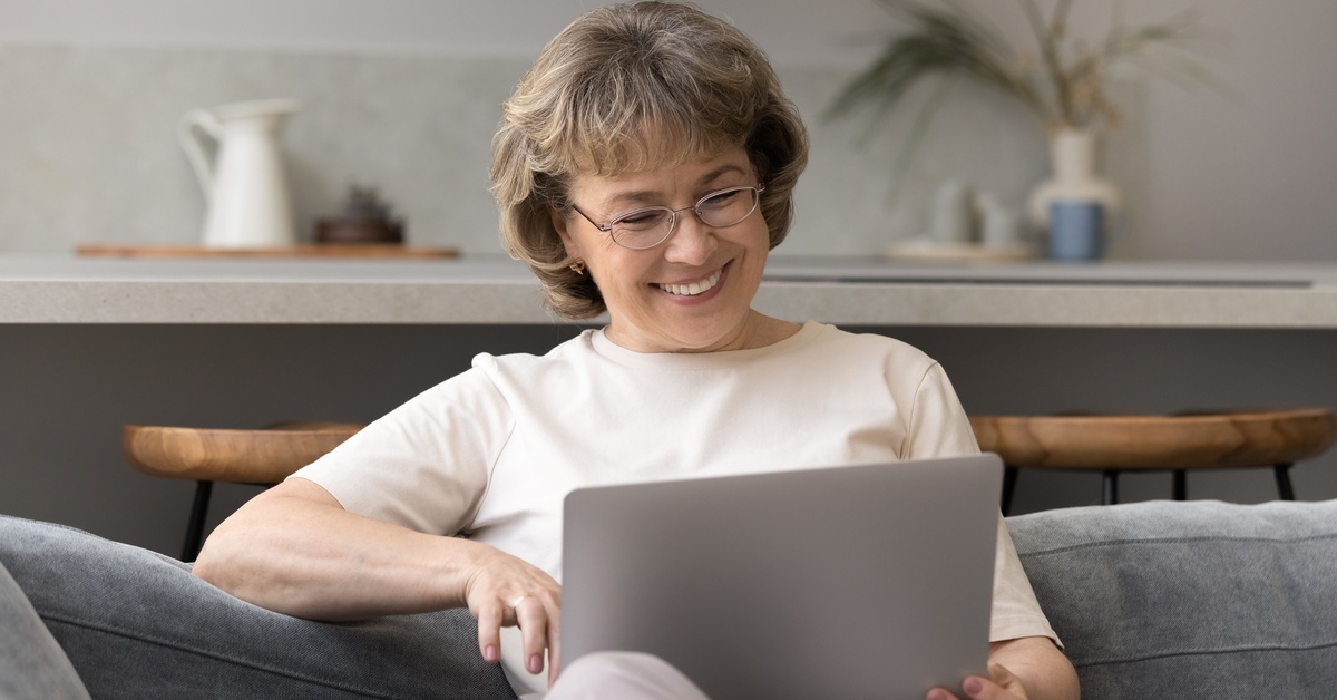 A woman wearing glasses and a white T-shirt smiles while sitting on a couch and staring at a laptop.