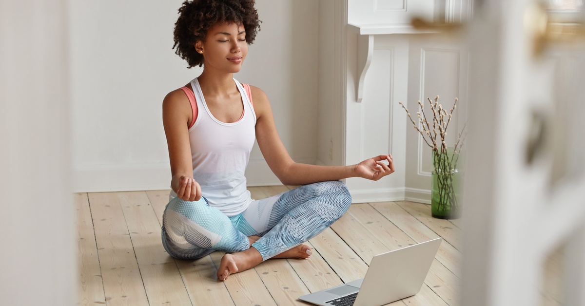 A young woman wearing a white tank top and patterned leggings sits on the floor with her legs crossed while meditating.
