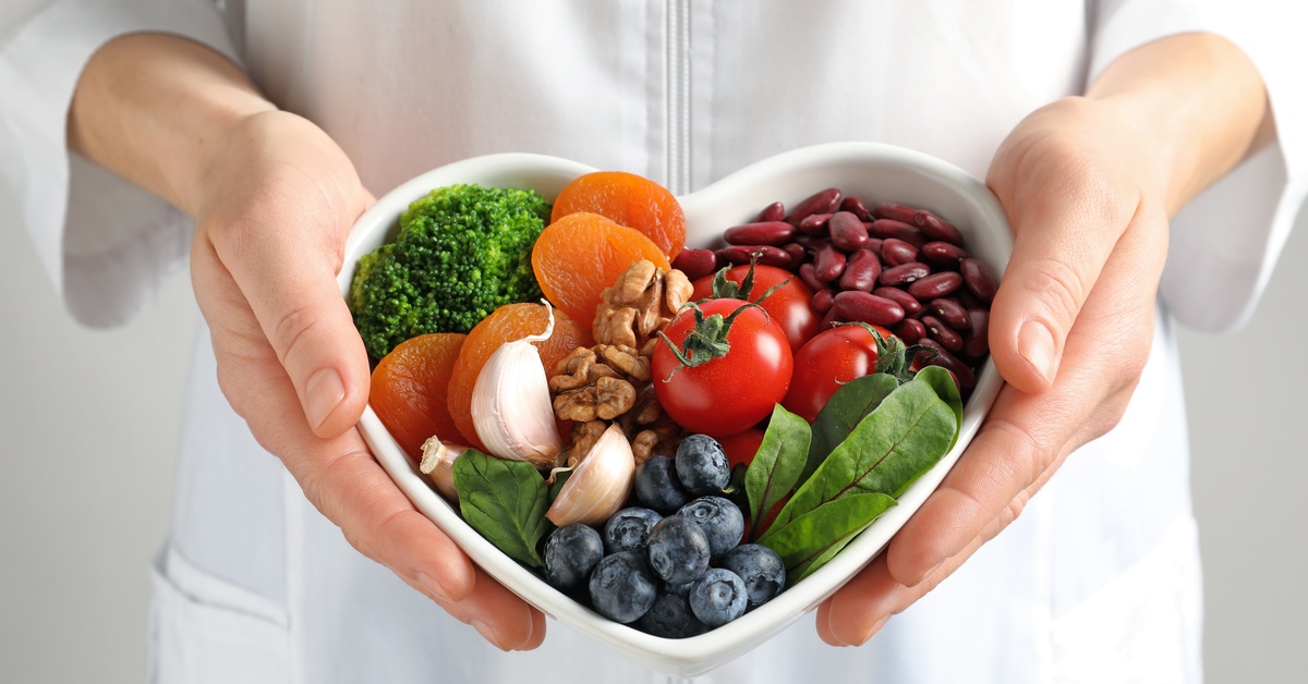 A person wearing a white jacket holds a heart-shaped bowl filled with heart-healthy foods, including broccoli and blueberries.