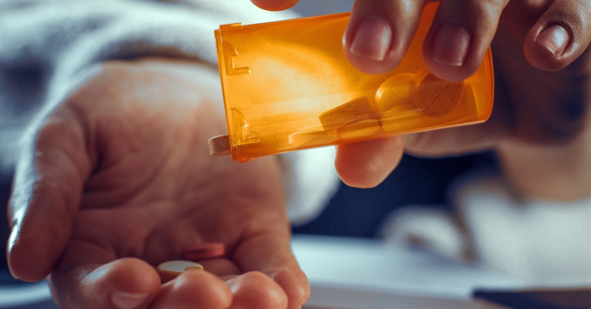 A person wearing a white shirt holds an orange bottle and pours out white pills into the palm of their hand.