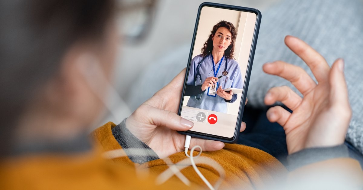A woman holds a phone and speaks to a doctor via a video call. The doctor has a clipboard and is talking to the woman.
