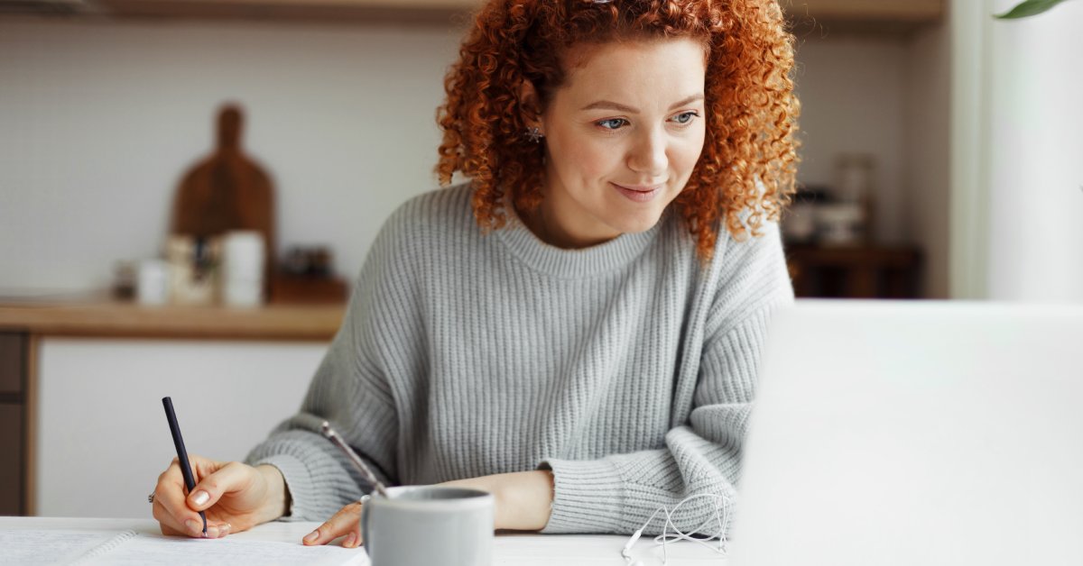 A young woman with glasses on top of her red, curly hair writes on a notepad while staring at a laptop screen.