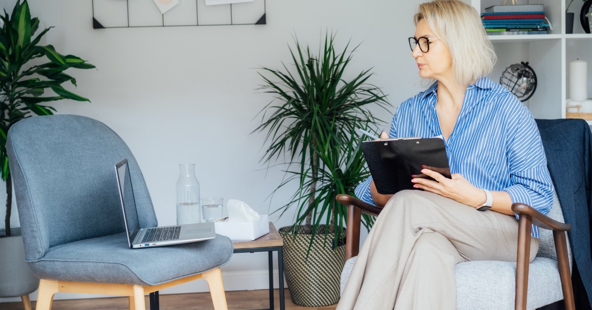 A woman wearing a blue striped shirt and khaki pants holds a clipboard while staring at a laptop screen in another chair.