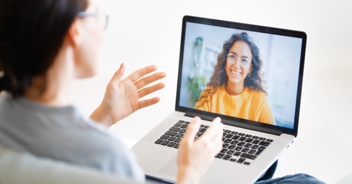 A young woman wearing an orange sweater smiles while communicating with another woman through a laptop screen.