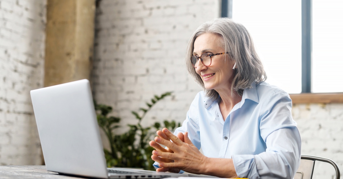 An older woman with grey hair smiles and places her hands together. She wears AirPods and looks directly at a laptop screen.