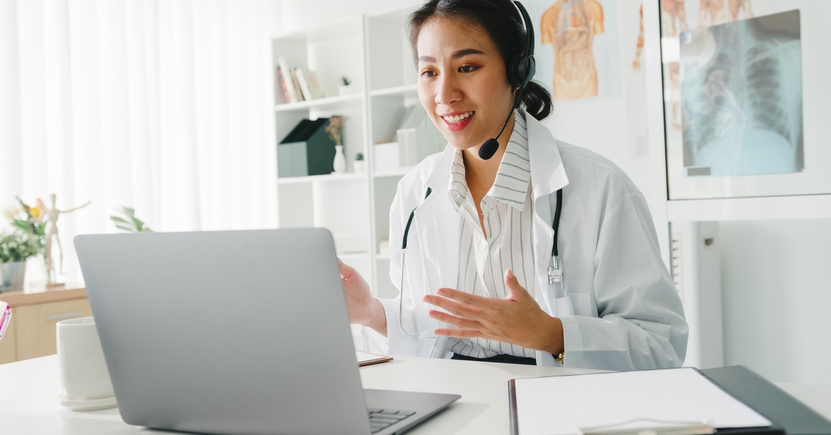 An Asian doctor wearing a white lab coat and a striped, white shirt speaks into a microphone while looking at a laptop screen.