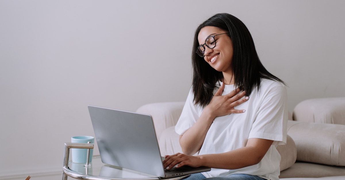 A young woman wearing glasses and a white t-shirt smiles and places her right hand on her chest while staring at a laptop screen.