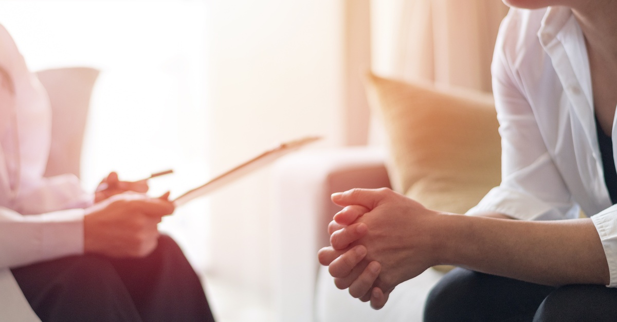 A young woman leans forward with her hands clasped together during a therapy session with a medical professional.