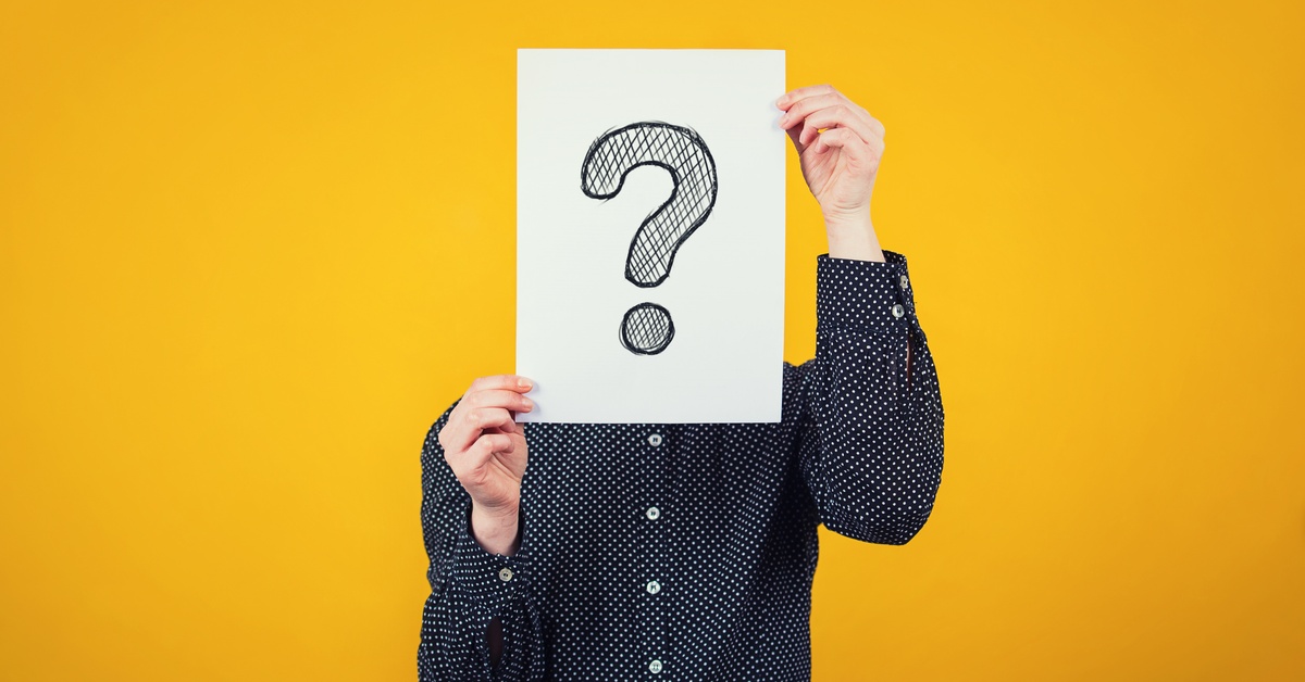 A person wearing a polka-dot button-down shirt holds a large sheet of white paper with a question mark on the front.