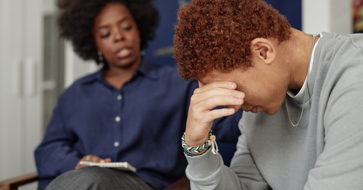 A young man wearing a grey sweatshirt appears upset as he receives support from a Black woman therapist.