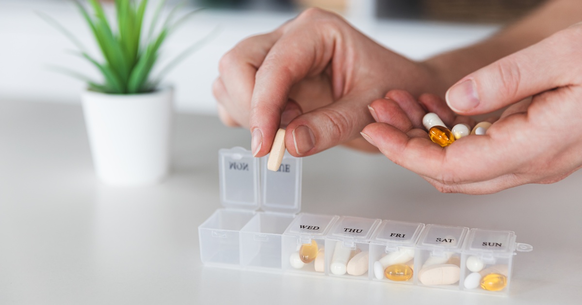 A woman uses her hands to place a colorful assortment of pills into a medical pill box labeled with the days of the week.