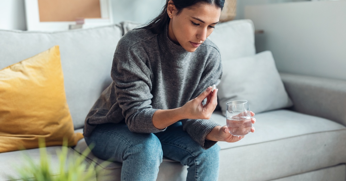 A young woman wearing a grey sweater sits on a couch while holding a glass of water and a pill.