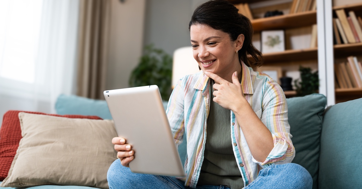 A woman with long, dark hair sits cross-legged on a couch and smiles while holding a tablet device.