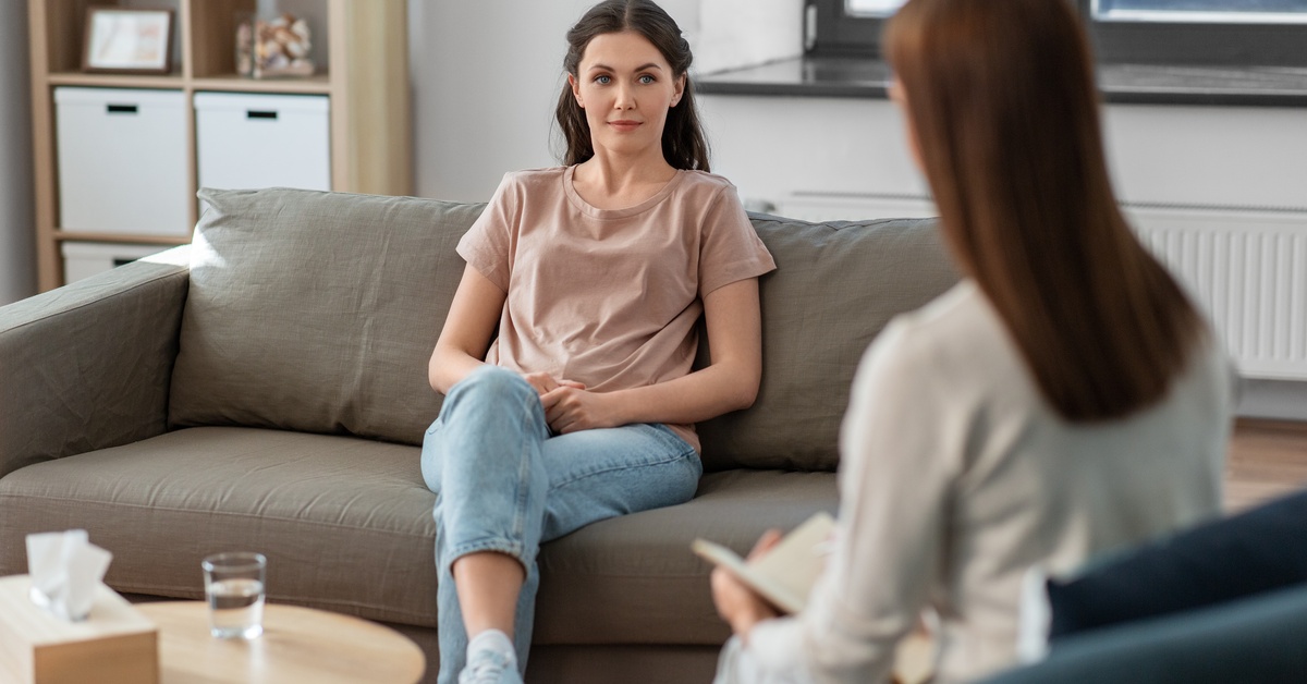 A young woman with long, dark hair sits on a couch and listens to a medical professional during a therapy session.
