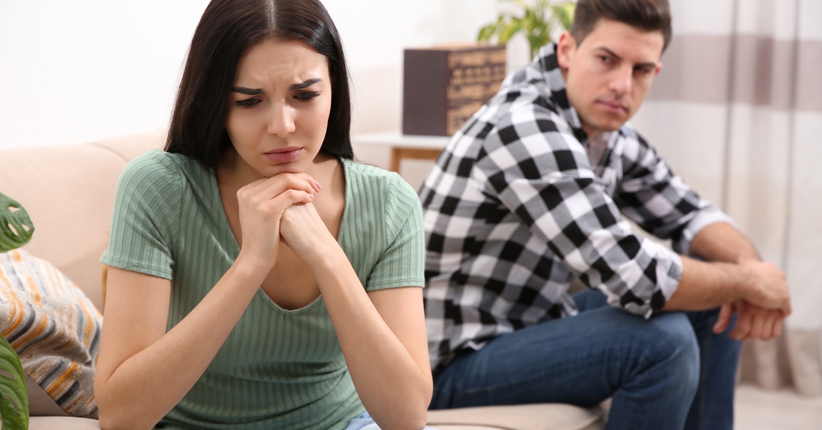 A young woman sits on the edge of a couch with a sad look on her face. A man sits on the other side and stares angrily at her.