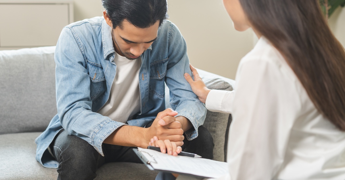 A young man looks sad during a session with a mental health therapist. The therapist touches his arm and attempts to console him.