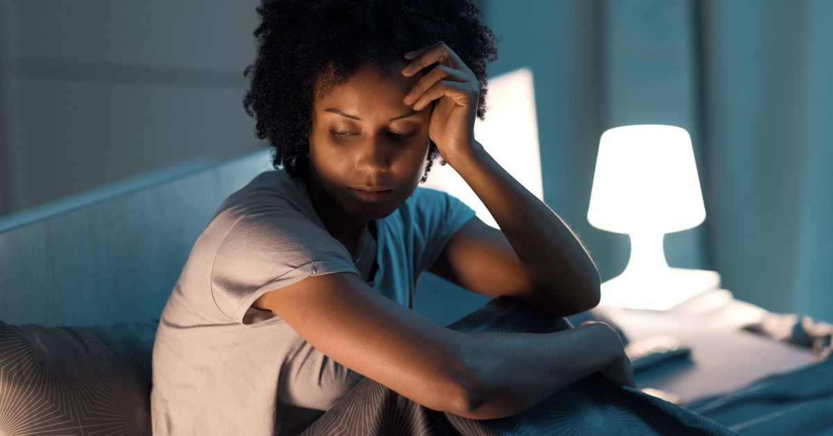 A young woman looks stressed as she sits in bed with her hand resting on her face. She’s in a dark room with a bright lamp.