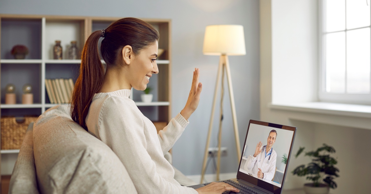 A woman sitting in a chair with a laptop on her lap, engaged in a video call with a doctor. They exchange waves.
