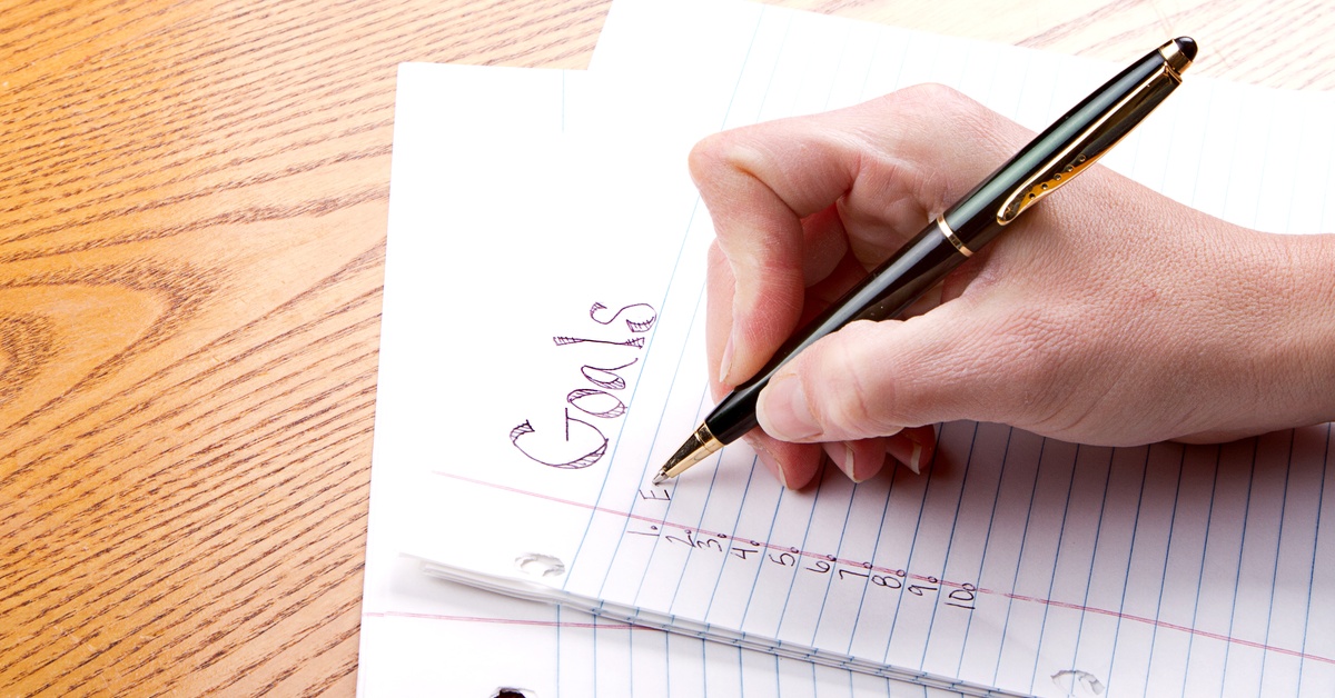 A hand holding a pen writing goals on a numbered list atop a stack of lined paper on a wooden surface.