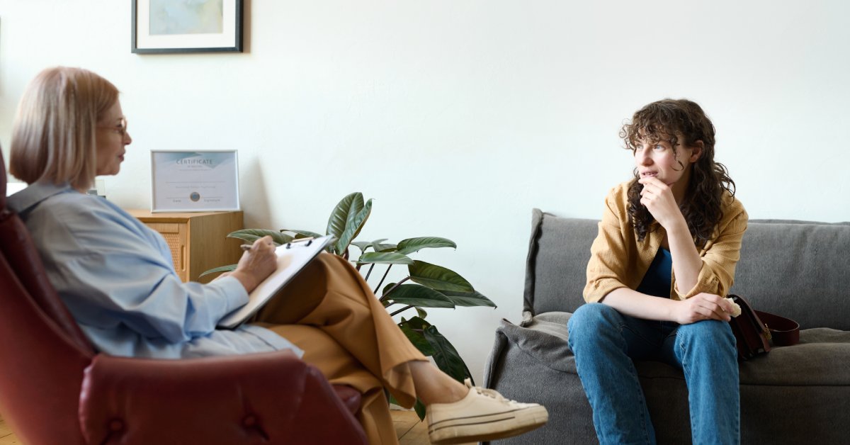 A young woman with curly hair sits on a sofa, her hand resting on her face. She speaks with a woman holding a clipboard.