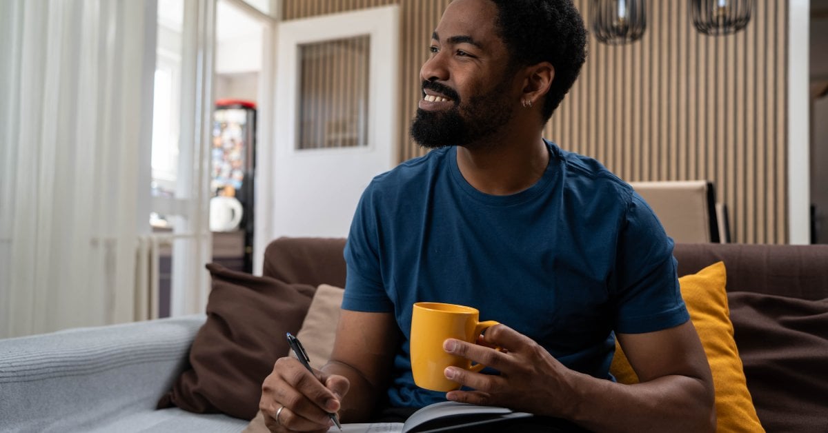 A young black man with a beard sits on a couch in a modern home. He smiles while holding a coffee mug and writing in a notebook.