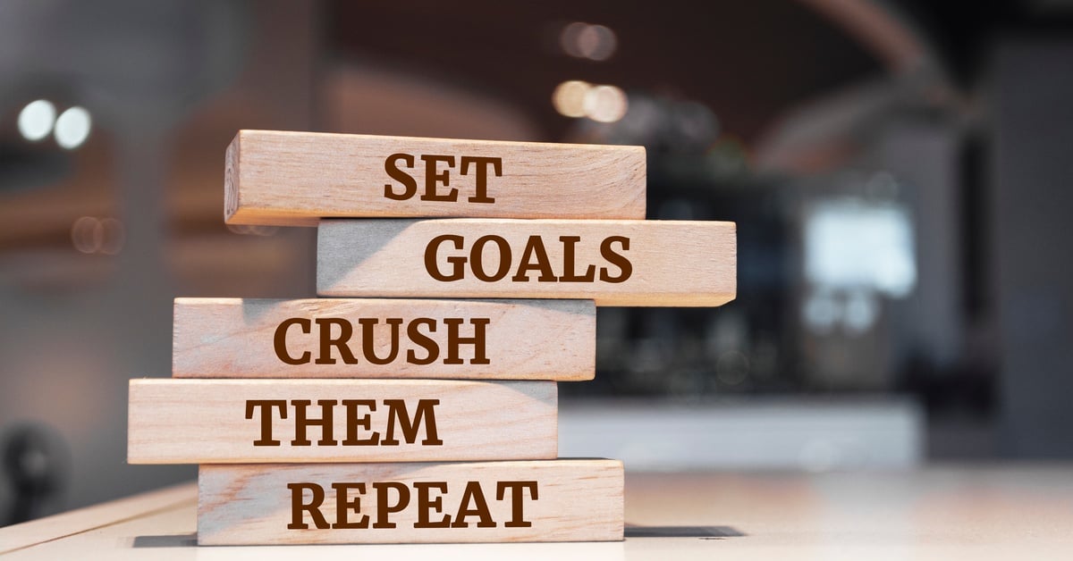 Five wooden blocks stand on top of each other. The blocks have words on them that say, “Set Goals Crush Them Repeat.”