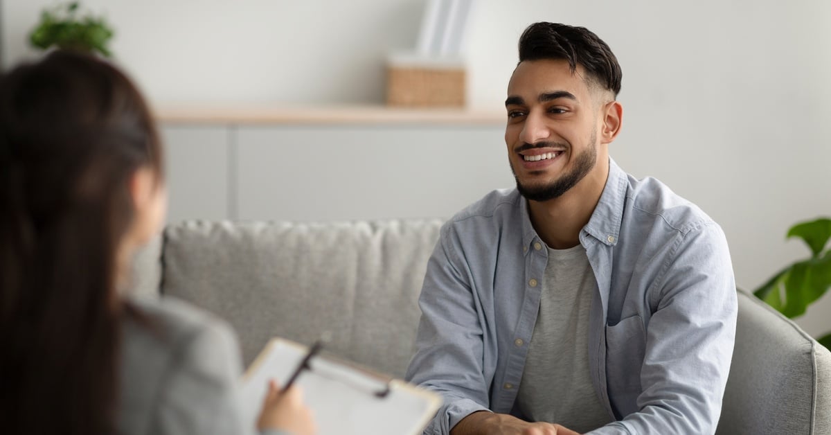 A young, Middle Eastern man smiles while sitting on a couch. He’s sitting directly across from a therapist holding a clipboard.