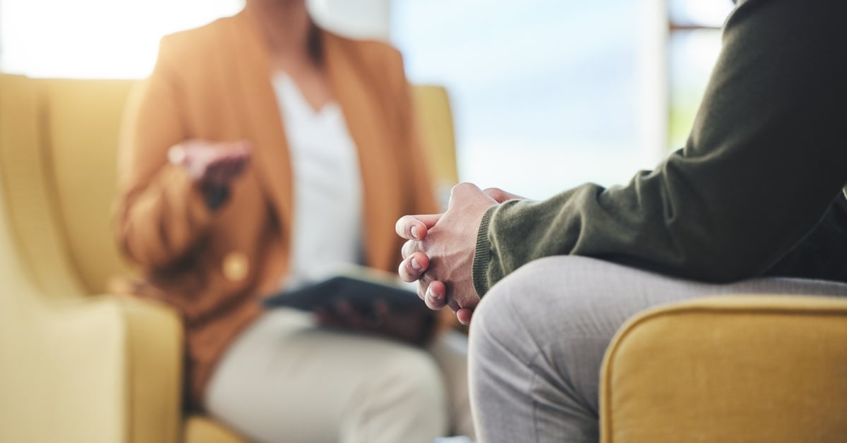 A woman therapist and a patient sit across from one another. The woman holds a clipboard, and the man clasps his hands together.