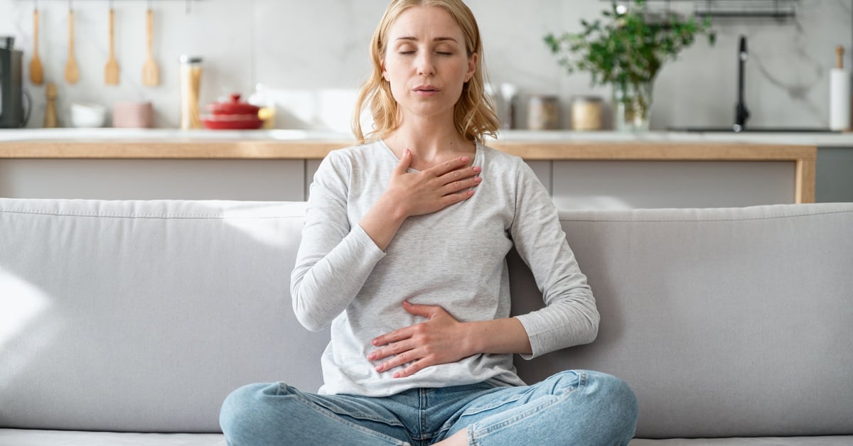 A woman sits on a couch, placing a hand on her chest and another on her stomach. She closes her eyes and focuses on her breathing.
