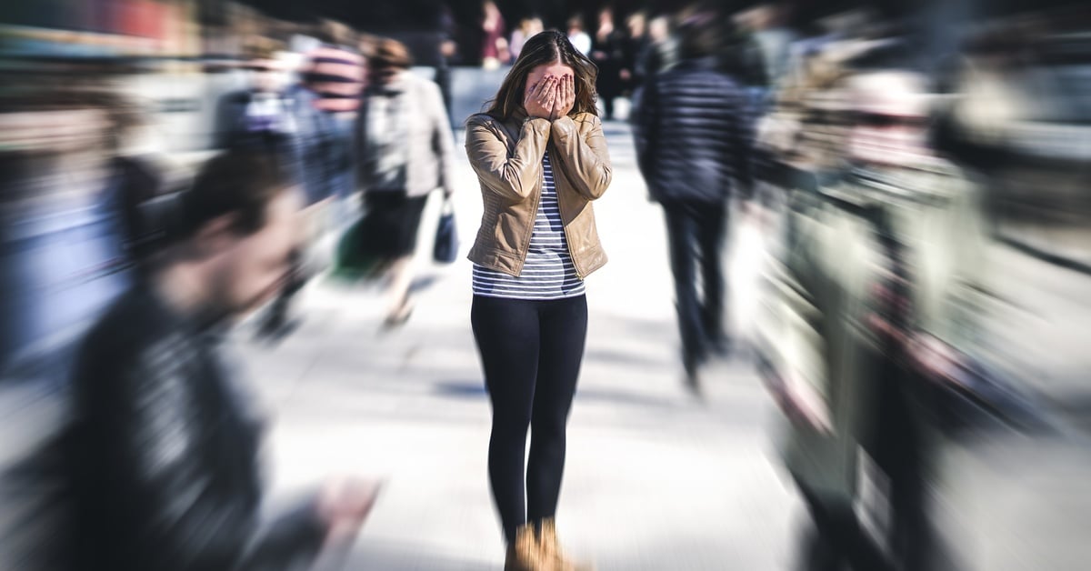 A young woman wearing a brown leather jacket hides her face in her hands in public. She appears to have an anxiety attack.