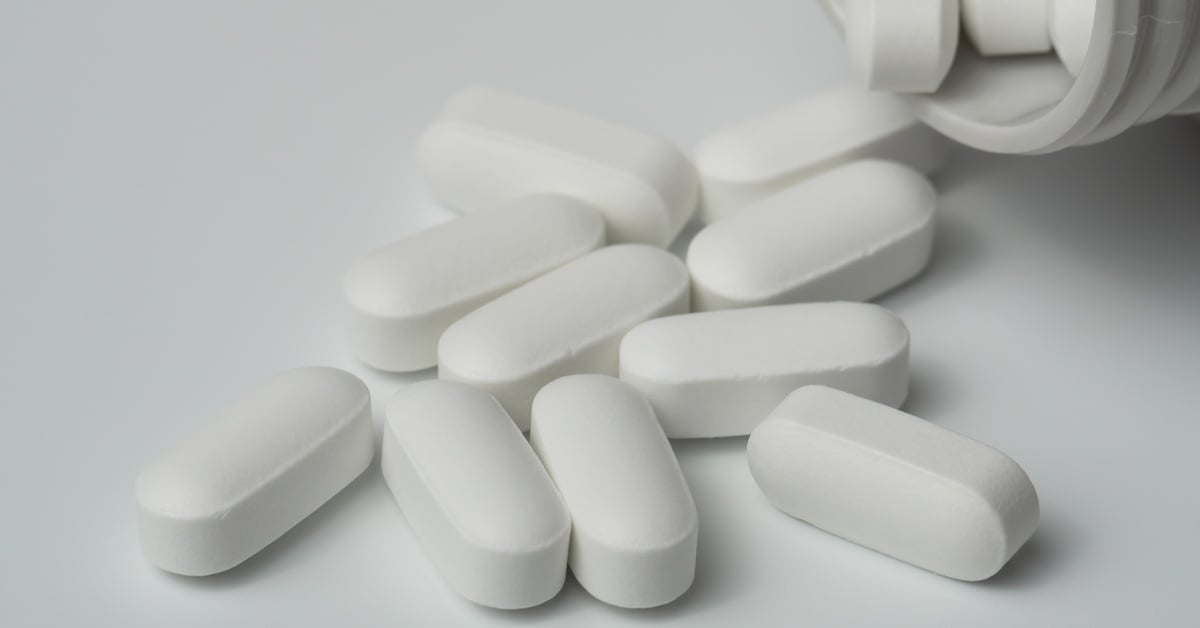 A bunch of white pills spill out of a bottle and land on a light grey surface. The pills have a capsule shape.