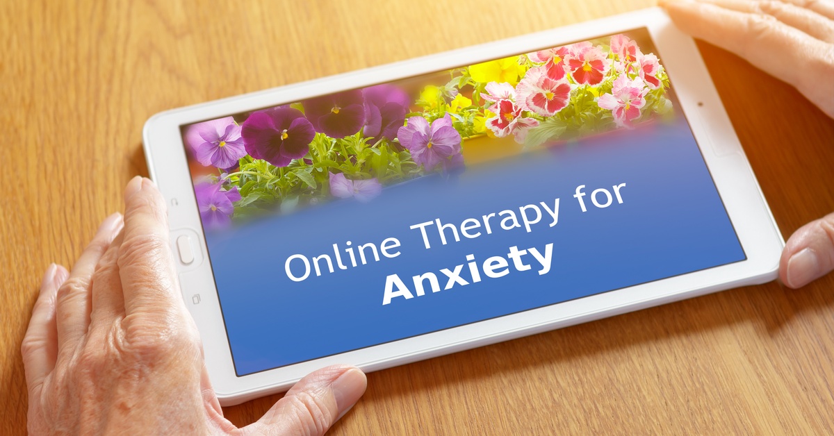 Hands hold a tablet on a wooden table. The tablet displays flowers and the words "Online Therapy for Anxiety" on the screen.
