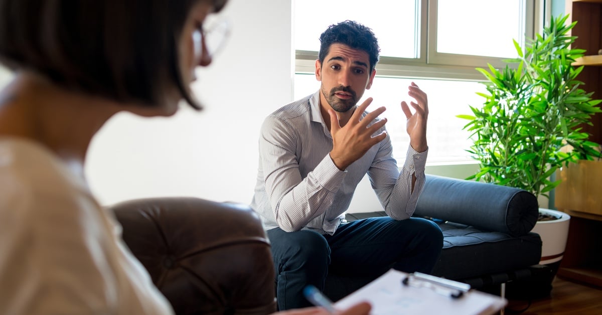 A man speaks with expressive gestures during a therapy session, while a therapist takes notes on a clipboard.