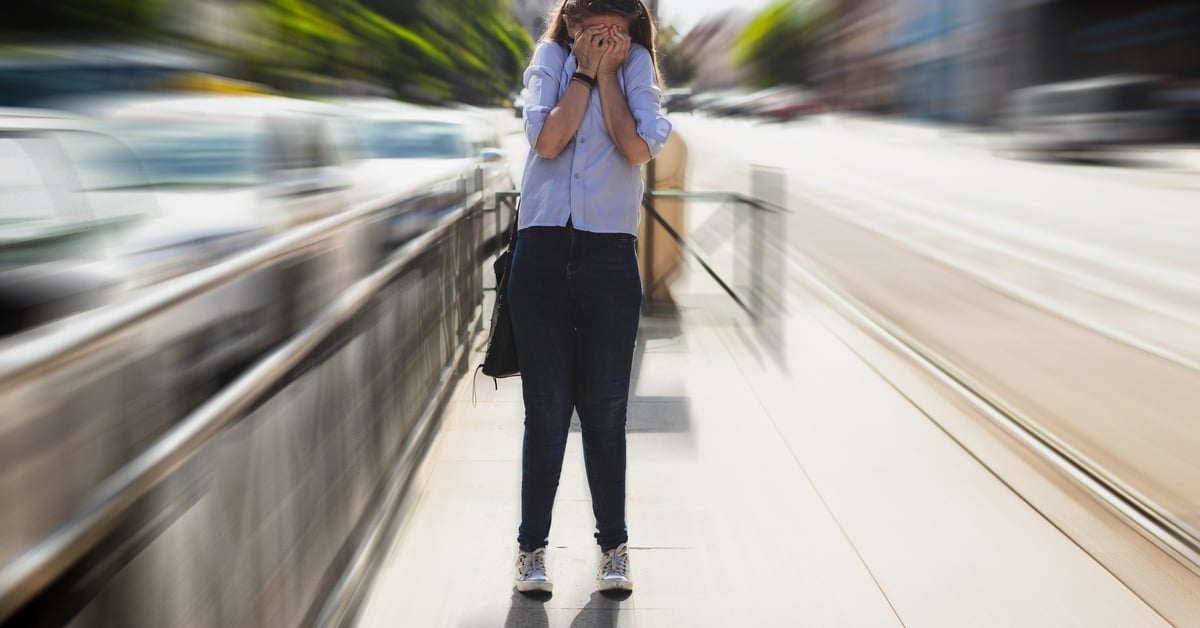 A woman standing on a city street covers her face with her hands, showing visible signs of anxiety and distress in public.