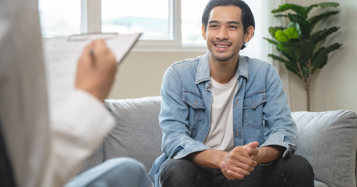 A smiling man in casual clothes sits on a couch and talks with a therapist, receiving support for managing anxiety.