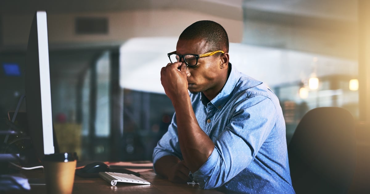 A man in glasses sits at a desk, holding his face in stress while working on a computer in a dimly lit office.