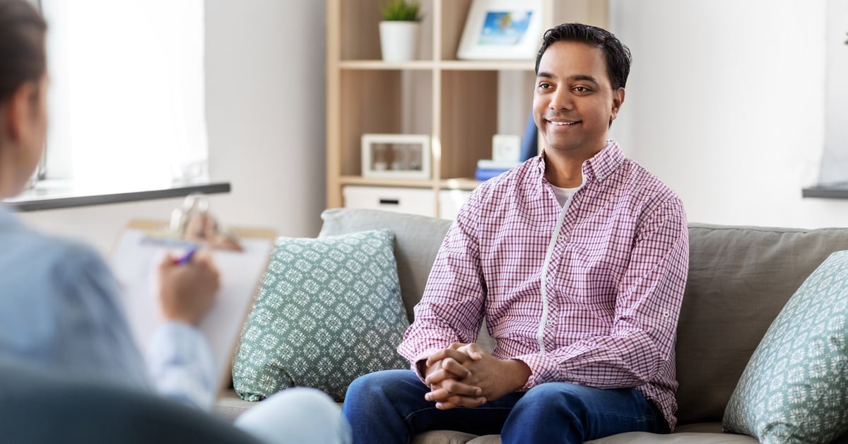 A male patient sits on a couch, smiling and looking toward a therapist who takes notes on a clipboard.